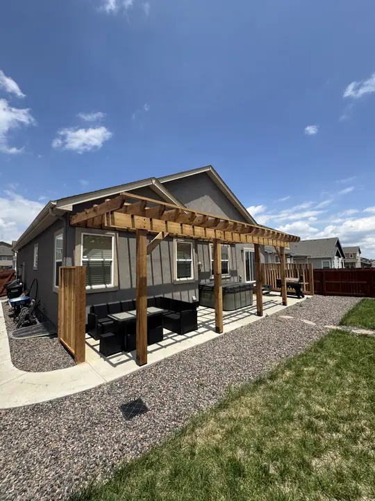 A long cedar pergola attached along the back of a gray house, over a patio with outdoor seating