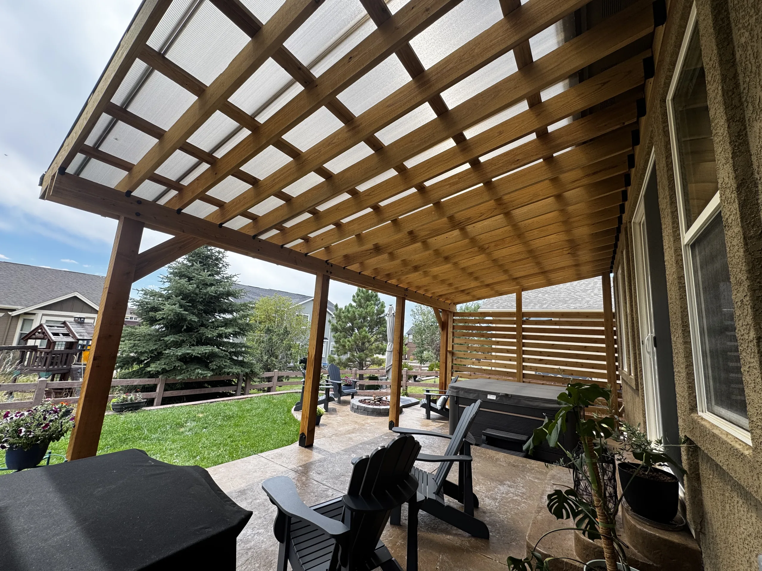 Looking out from under a covered cedar pergola along the side of a house, with chairs, a hot tub and a slatted privacy screen